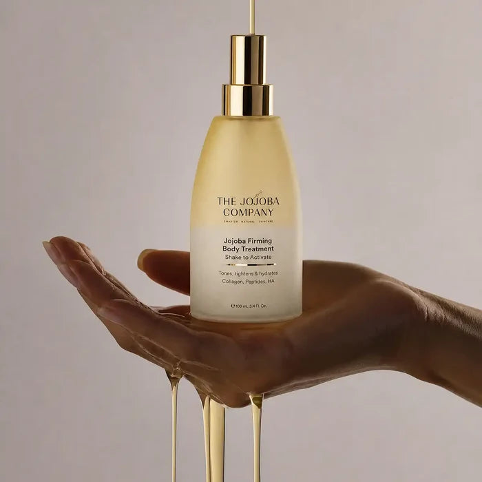 A hand holds a frosted bottle of The Jojoba Company Jojoba Firming Body Treatment, with golden honey-like product dripping from the bottle and around the hand against a soft beige background.