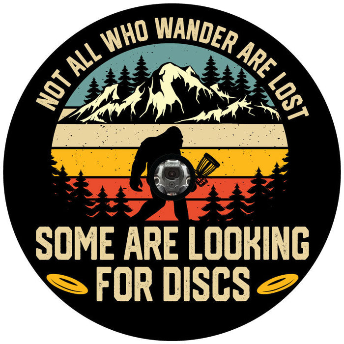 A funny spare tire cover design of sasquatch walking through the wilderness holding a disc golf basket and the saying not all who wonder are lost some are looking for discs on black background with a JL back up camera