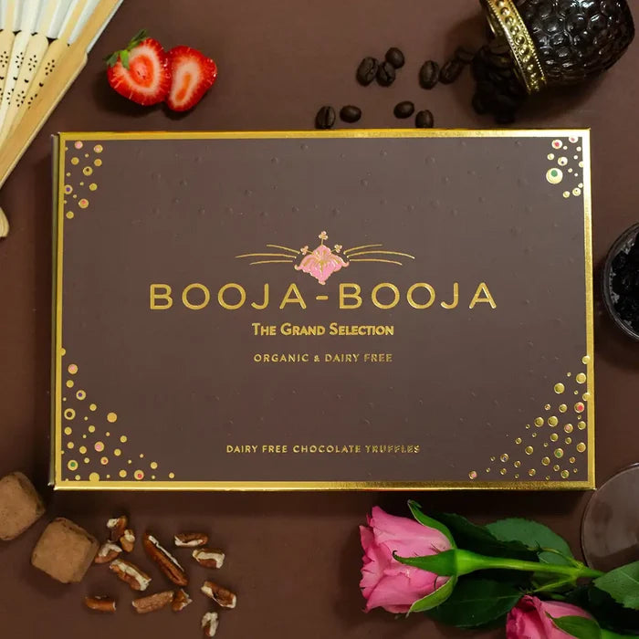 A rectangular chocolate truffle box in dark brown with gold lettering and decorative gold dots, styled on a brown surface with strawberries, coffee beans, pink roses, and chocolate pieces around it.